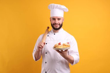 Üniformalı mutlu pastacı elinde çilekli profiterol ve turuncu arka planda merdane tutuyor.