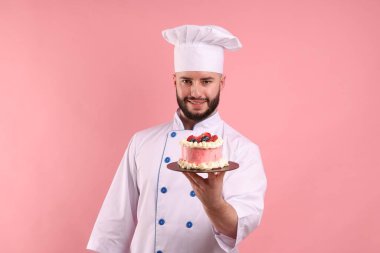 Üniformalı mutlu pastacı pembe arka planda böğürtlenli pasta tutuyor.