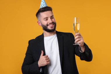 Doğum günün kutlu olsun. Parti şapkası ve şampanyası olan yakışıklı adam turuncu arka planda baş parmak gösteriyor.