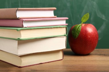 Tahtanın yanındaki ahşap masada farklı kitaplar ve kırmızı elma, yakın plan.