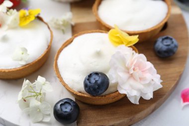 Lezzetli tatlı. Kremalı tartlar, böğürtlenler ve çiçekler beyaz döşemeli masada, yakın plan.