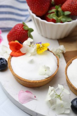 Lezzetli tatlı. Kremalı tartlar, böğürtlenler ve çiçekler beyaz döşemeli masada, yakın plan.