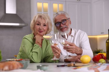Yaşlı çift mutfakta yemek pişirirken beyaz şarap içiyor.