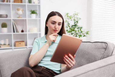 Evdeki koltukta modern tablet kullanan genç bir kadın.