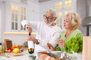 Yaşlı bir adam mutfakta karısıyla yemek pişirirken şarap şişesini açıyor.