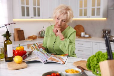 Mutfaktaki mermer masada gözlüklü yaşlı kadın dergisi okuyor.