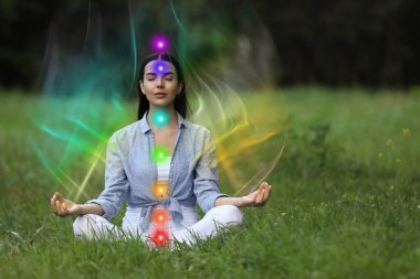 Lotus 'ta meditasyon yapan kadın yeşil çimlerde poz veriyor. Etrafında gökkuşağı dalgaları gibi görünen bir hava. Yedi çakra parlıyor