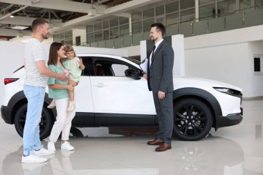 Gülümseyen satıcı elinde dosyayla salonda aileye yeni bir araba gösteriyor.