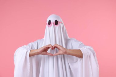 Şirin hayalet. Güneş gözlüklü beyaz çarşaflı adam pembe arka planda kalp hareketi yapıyor.