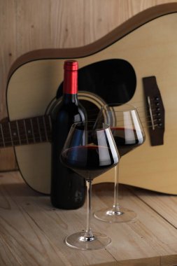 Akustik gitar, şişe ve kırmızı şarap kadehleri ahşap masada.