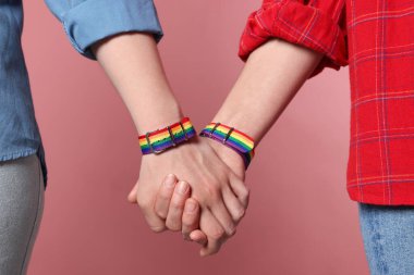 LGBT konsepti. Gökkuşağı bileklikli kadınlar pembe arka planda el ele tutuşuyorlar, yakın plan.