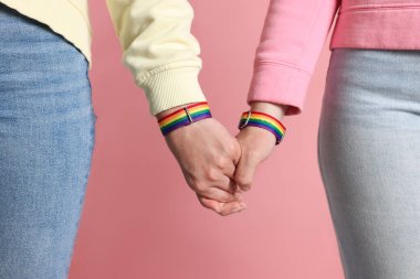 LGBT konsepti. Gökkuşağı bileklikli kadınlar pembe arka planda el ele tutuşuyorlar, yakın plan.