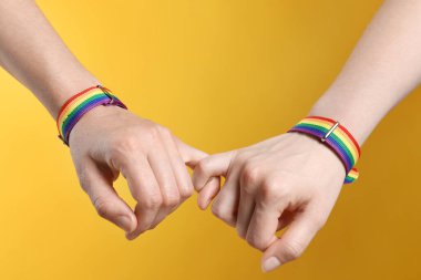 LGBT konsepti. Gökkuşağı bileklikli kadınlar sarı arka planda el ele tutuşuyorlar, yakın plan.