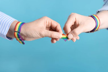 LGBT konsepti. Açık mavi arka planda kalp şeklinde iğnesi olan gökkuşağı bileklikli kadınlar, yakın plan