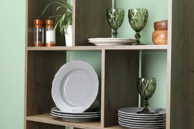 İçerideki yeşil duvarın yanında farklı bulaşık takımları, gözlük ve çiçek dekoru olan raf ünitesi, yakın plan.