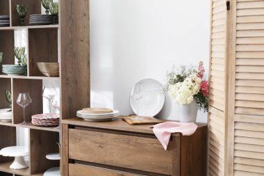 Beyaz duvarın yanında farklı bulaşık takımları, gözlük ve çiçek dekoru olan raf ünitesi.