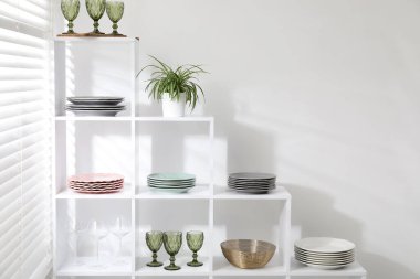 Beyaz duvarın yanında farklı bulaşık takımları, gözlük ve çiçek dekoru olan raf ünitesi.