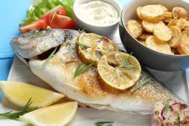 Hafif mavi ahşap masada sebze ve sosla servis edilen kızarmış dorado balığı, yakın plan. Ev yapımı deniz ürünleri yemeği.