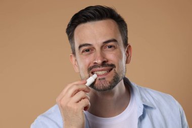 Gülümseyen adam açık kahverengi arka planda dudaklarına merhem sürüyor.