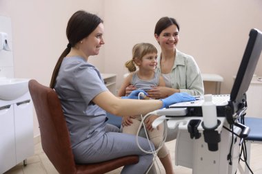 Sonograf küçük bir kızın karın ultrasonu muayenesini yönetiyor. Anne ve kızının klinikte doktorla randevusu var.