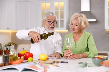Yaşlı çift mutfakta yemek pişirirken beyaz şarap içiyor.