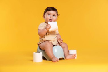 Küçük şirin bebek tuvalette oturuyor ve tuvalet kağıdı ruloları turuncu arka planda.