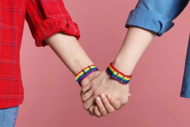 LGBT konsepti. Gökkuşağı bileklikli kadınlar pembe arka planda el ele tutuşuyorlar, yakın plan.