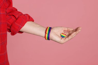 LGBT konsepti. Pembe arka planda kalp şeklinde iğnesi olan gökkuşağı bileklikli kadın, yakın plan.