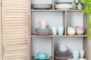 Beyaz duvarın yanında farklı bulaşık takımları, bardaklar, fincanlar ve çiçek dekoru olan raf ünitesi.