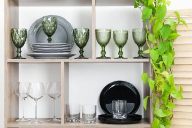 Beyaz duvarın yanında farklı bulaşık takımları, gözlük ve çiçek dekoru olan raf ünitesi.