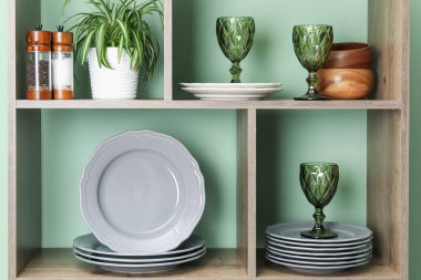 Farklı bulaşık takımları, gözlükler ve yeşil duvarın yanında çiçek dekoru olan raf ünitesi.