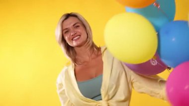 Sarı arka planda renkli balonlarla oynayan mutlu kadın.