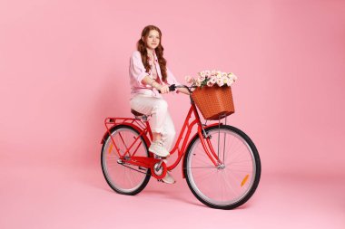 Güzel genç kız pembe arka planda bir sepet çiçekle bisiklete biniyor.