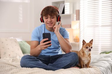 Evde akıllı telefon ve köpek kullanarak kulaklık takan gülümseyen genç bir çocuk.