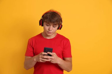 Kulaklıklı genç çocuk sarı arka planda akıllı telefon kullanıyor.