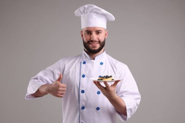 Üniformalı mutlu pastacı elinde yaban mersinli lezzetli bir tart ve açık gri arka planda baş parmak gösteriyor.
