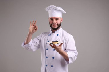 Üniformalı mutlu pastacı elinde yaban mersinli lezzetli bir tart tutuyor ve açık gri arka planda güzel bir jest yapıyor.