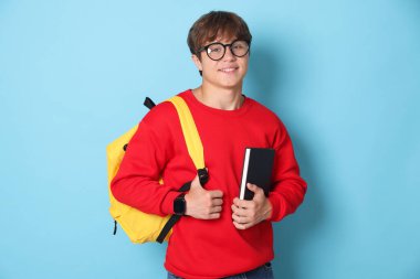 Sırt çantası açık mavi arka planda, gözlüklü, gülümseyen genç çocuk.