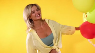 Sarı arka planda renkli balonlarla oynayan mutlu kadın.