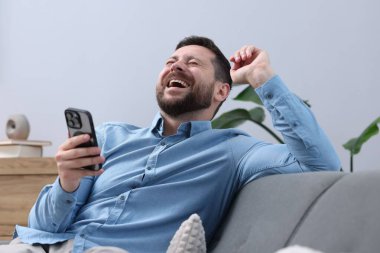 Akıllı telefonlu bir adam evdeki kanepede gülüyor.