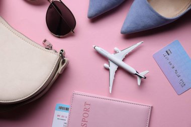 Pembe arka planda uçak modeli, pasaport, uçuş bileti, kredi kartı, ayakkabı ve aksesuarlar.