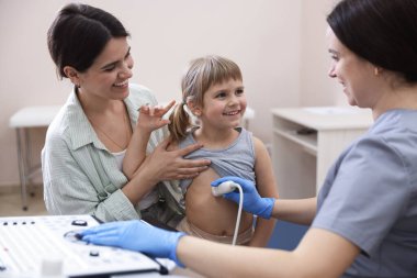 Sonograf küçük bir kızın karın ultrasonu muayenesini yönetiyor. Anne ve kızının klinikte doktorla randevusu var.