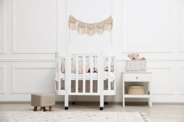 Rahat beşik, puf, komodin ve bebek odasındaki beyaz duvarın yanında oyuncaklar. İç tasarım