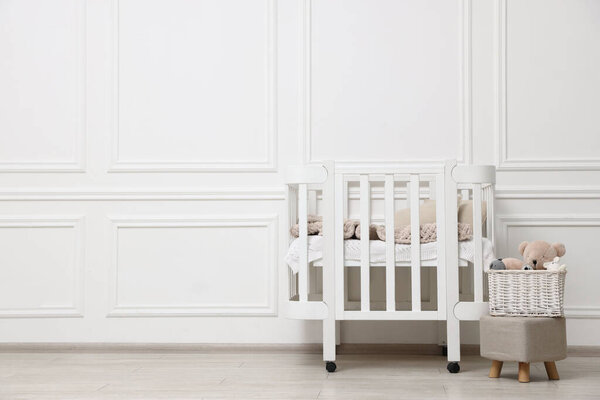 Comfortable crib, pouf and toys near white wall in baby's room, space for text. Interior design