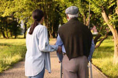 Bakıcı, dışarıda yürüyen çerçeve kullanan yaşlı bir adamı destekliyor.