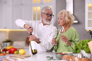 Yaşlı bir adam mutfakta karısıyla yemek pişirirken şarap şişesini açıyor.