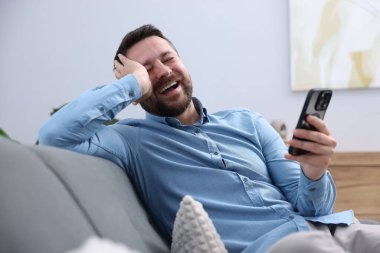 Akıllı telefonlu bir adam evdeki kanepede gülüyor.