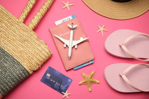 Uçak modeli, pasaport, uçuş bileti, kredi kartı ve pembe arka planda plaj aksesuarları.