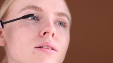 Genç bir kadın kahverengi arka planda kirpiklerine rimel sürüyor, yakın plan.