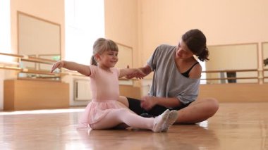 Gülümseyen bale öğretmeni dans dersinde küçük kızın yere uzanmasına yardım ediyor.